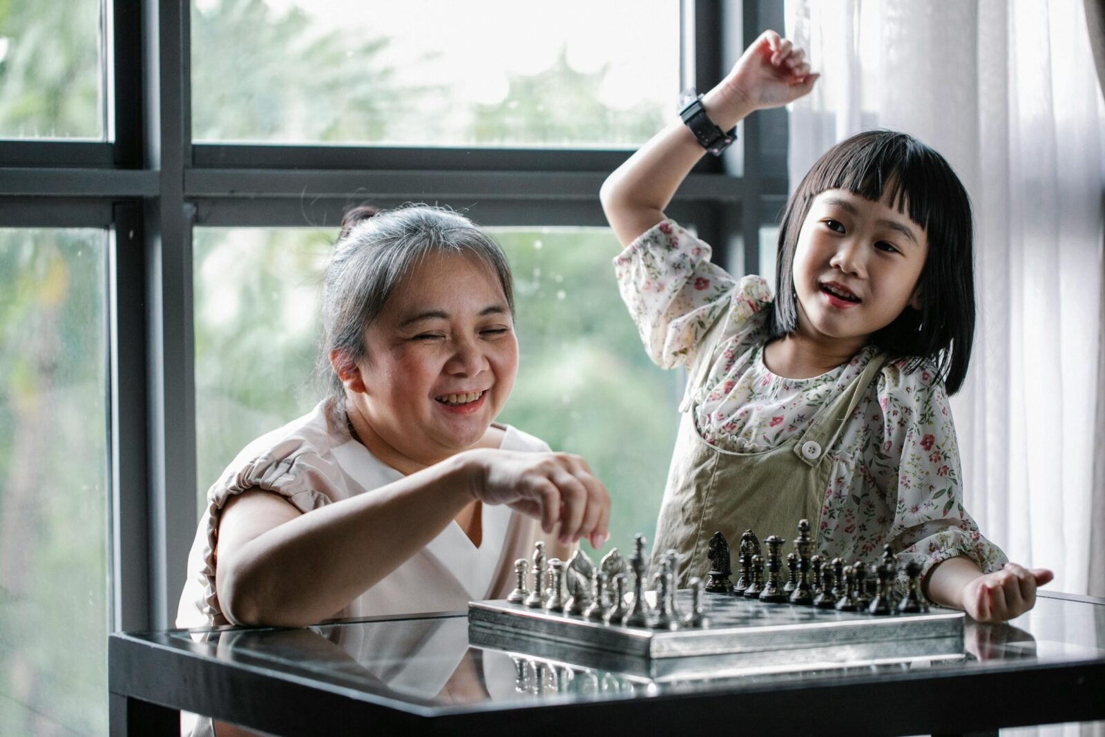 Happy little Asian girl with grandmother sitting at table and playing chess during weekend at home