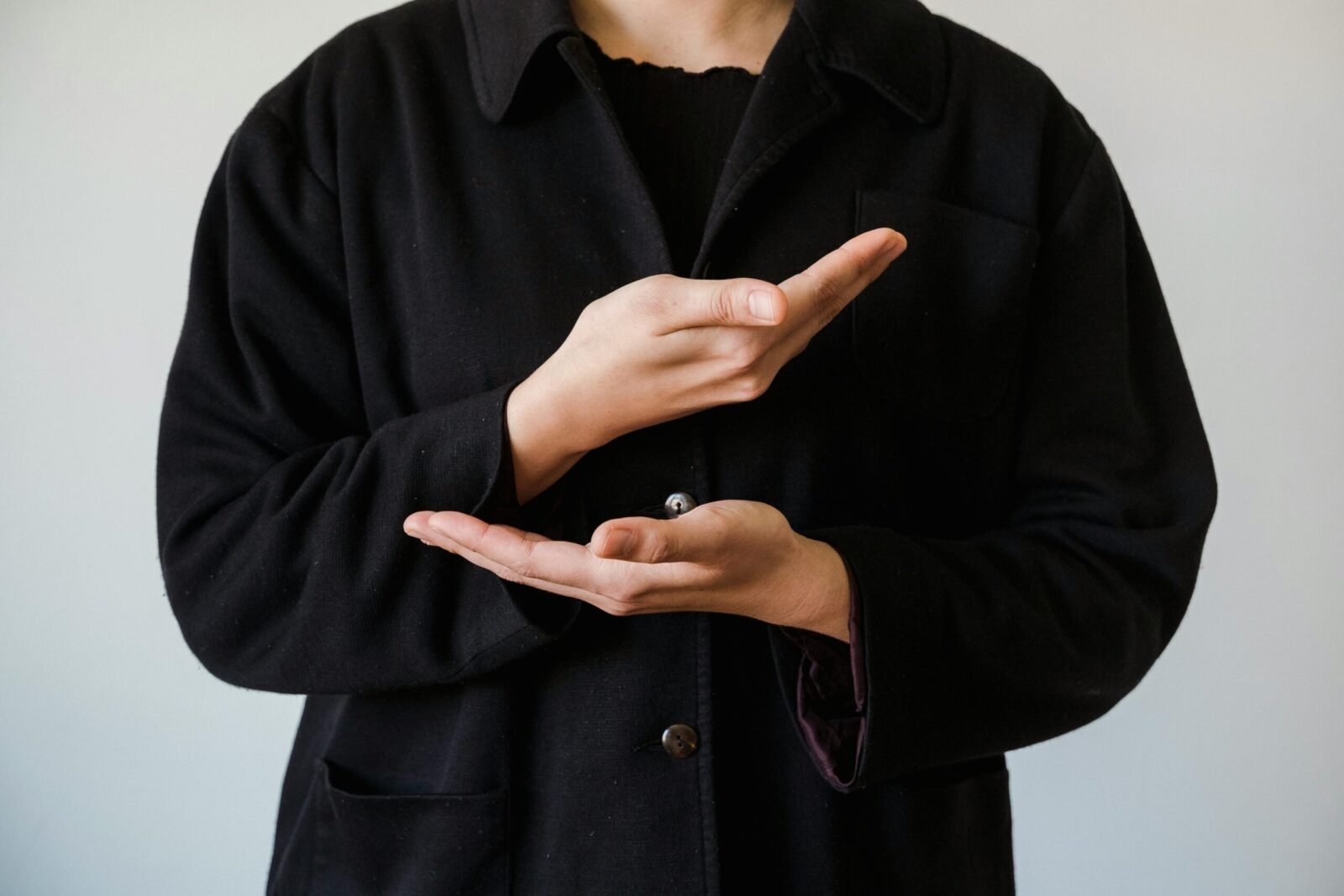 Close-up of a person gesturing with hands in a black coat, against a white background.